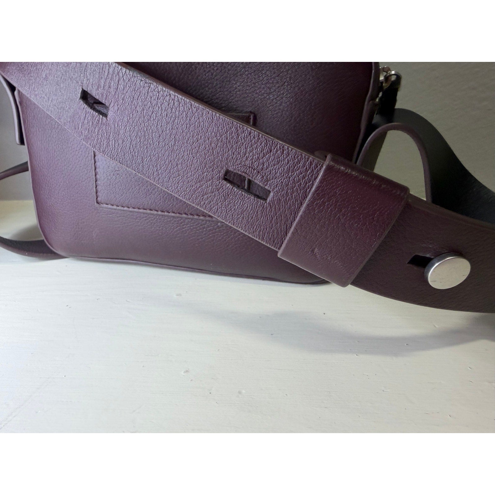 AllSaints women's burgundy pebbled leather camera crossbody bag with adjustable strap, showcasing a stylish handbag design.